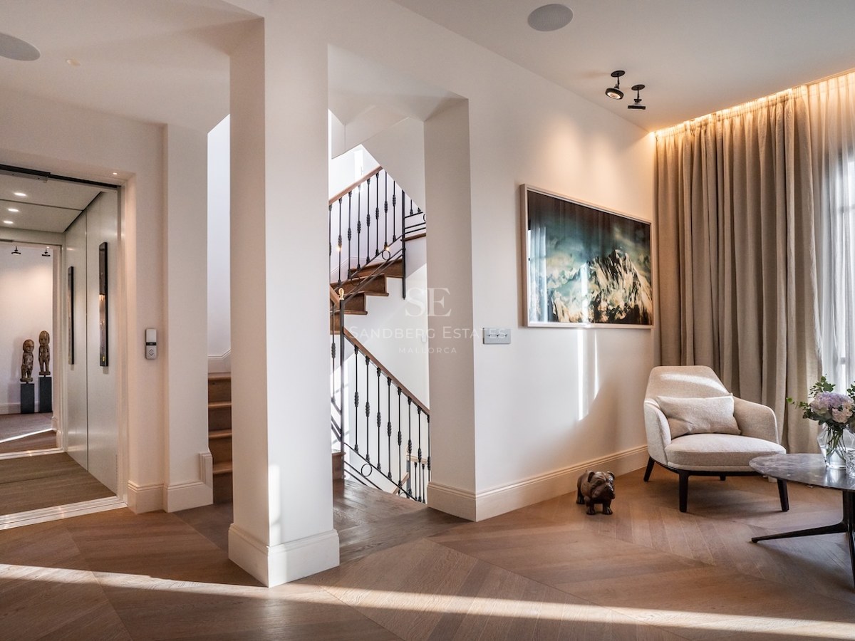 Pasillo de lujo con suelo de madera en espiga, ascensor privado, escalera y un sillón de diseño blanco.