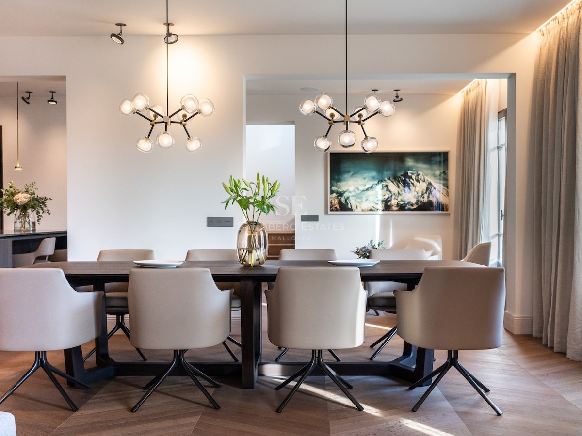 Comedor elegante con mesa de madera oscura, sillas beige, lámparas de esfera y cuadro de montaña.