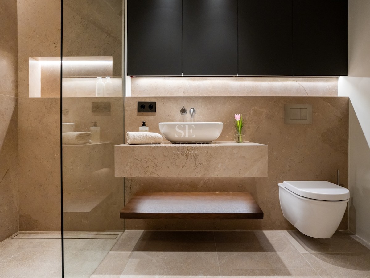 Baño moderno con paredes de piedra beige, lavabo flotante de mármol e iluminación LED integrada.