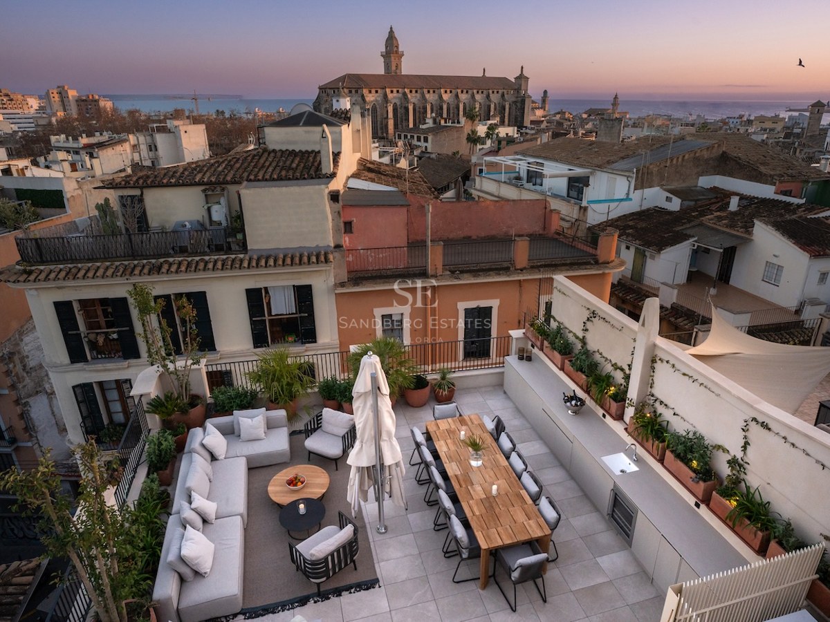 Elegante terraza en la azotea con sofás, mesa de comedor y cocina exterior frente a una catedral histórica al atardecer.