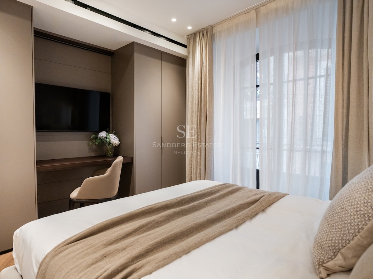 Dormitorio moderno con cama blanca, manta beige, escritorio integrado con TV y grandes ventanales con cortinas.