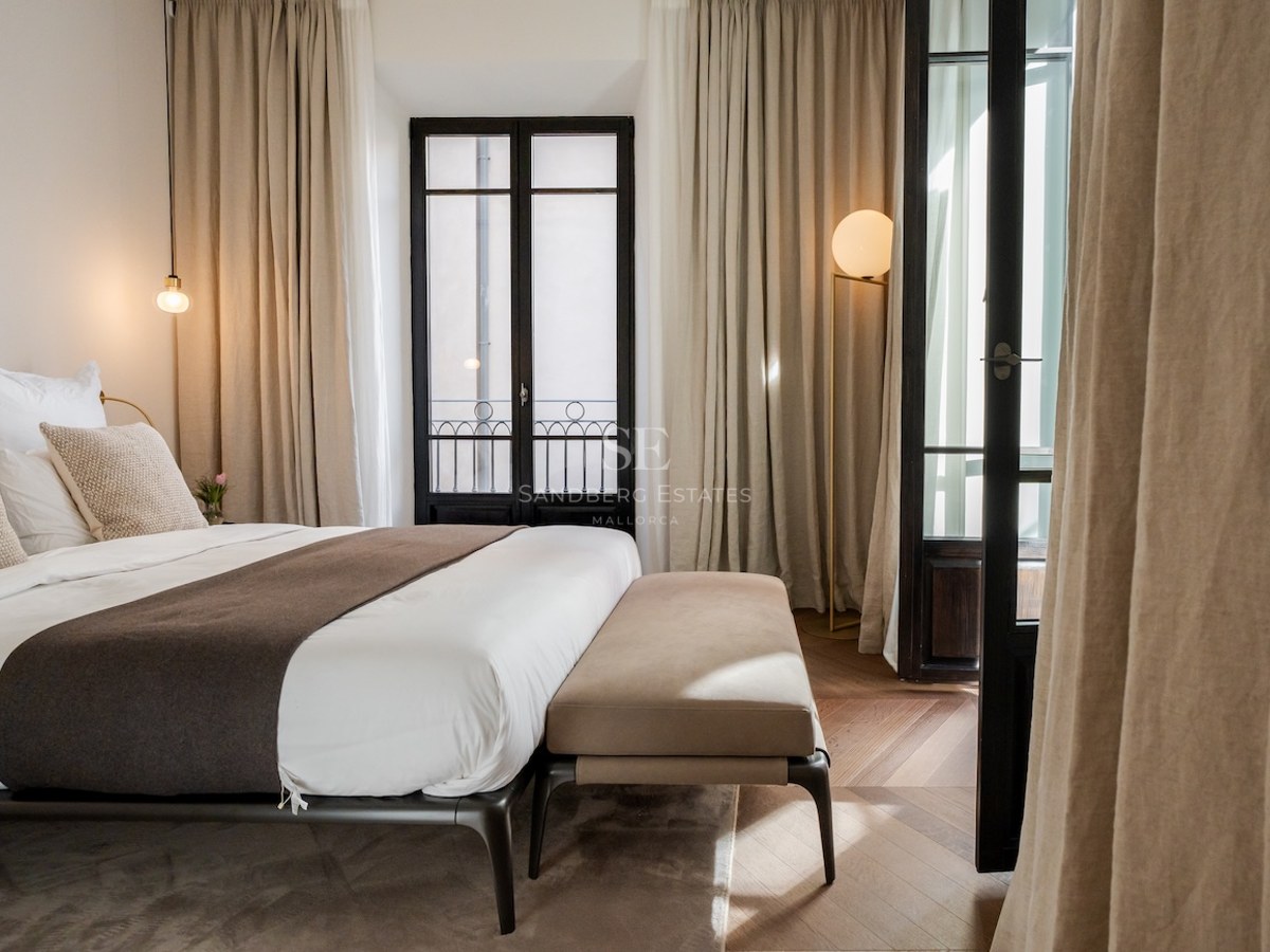 Dormitorio moderno con cama grande, cortinas de lino beige, suelo de madera en espiga y puertas de cristal negras.