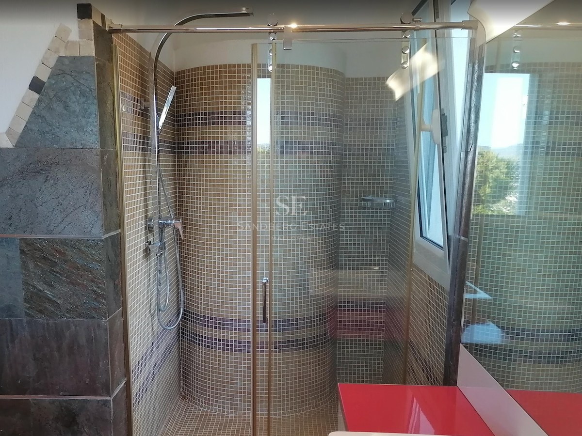 A walk-in glass shower enclosure featuring beige mosaic tiles, chrome fixtures, and a curved interior wall.