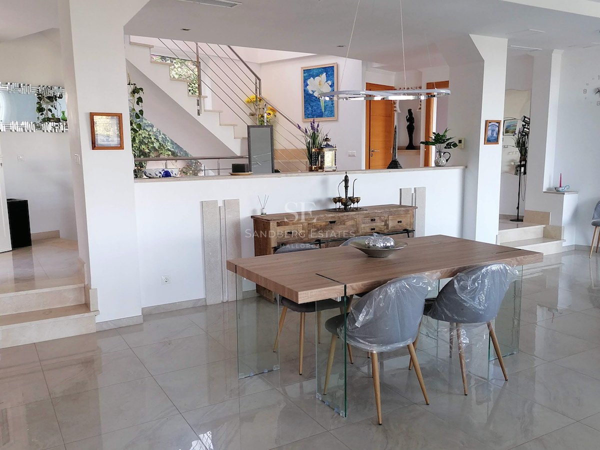 Modern dining room with wooden table, glass legs, marble floors, and stairs in the background.