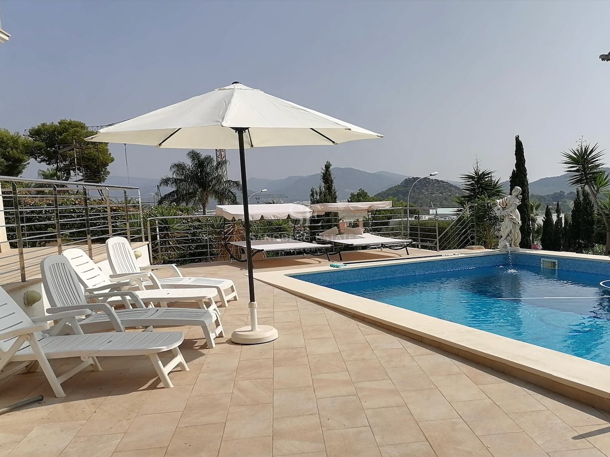 Sun-drenched pool area featuring white sun loungers, a large parasol, and a classic statue overlooking mountains.