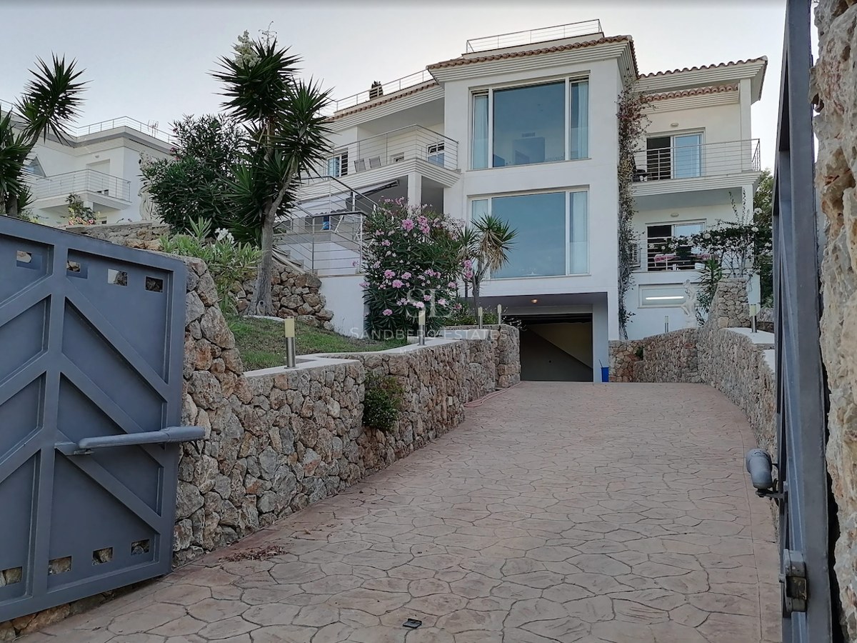 View of a multi-story white villa with large windows, seen from a gated driveway lined with natural stone walls.