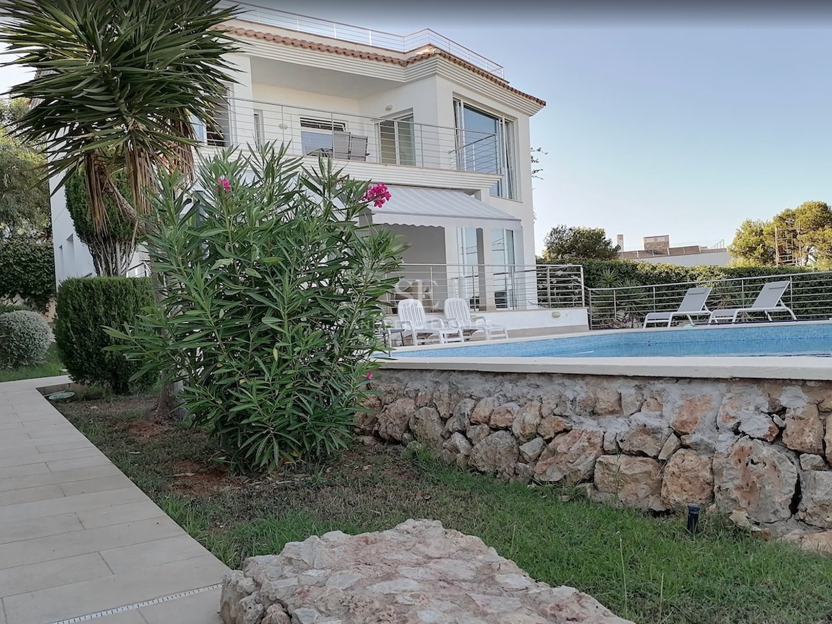 White two-story villa overlooking a turquoise pool with sun loungers and a natural stone garden wall.