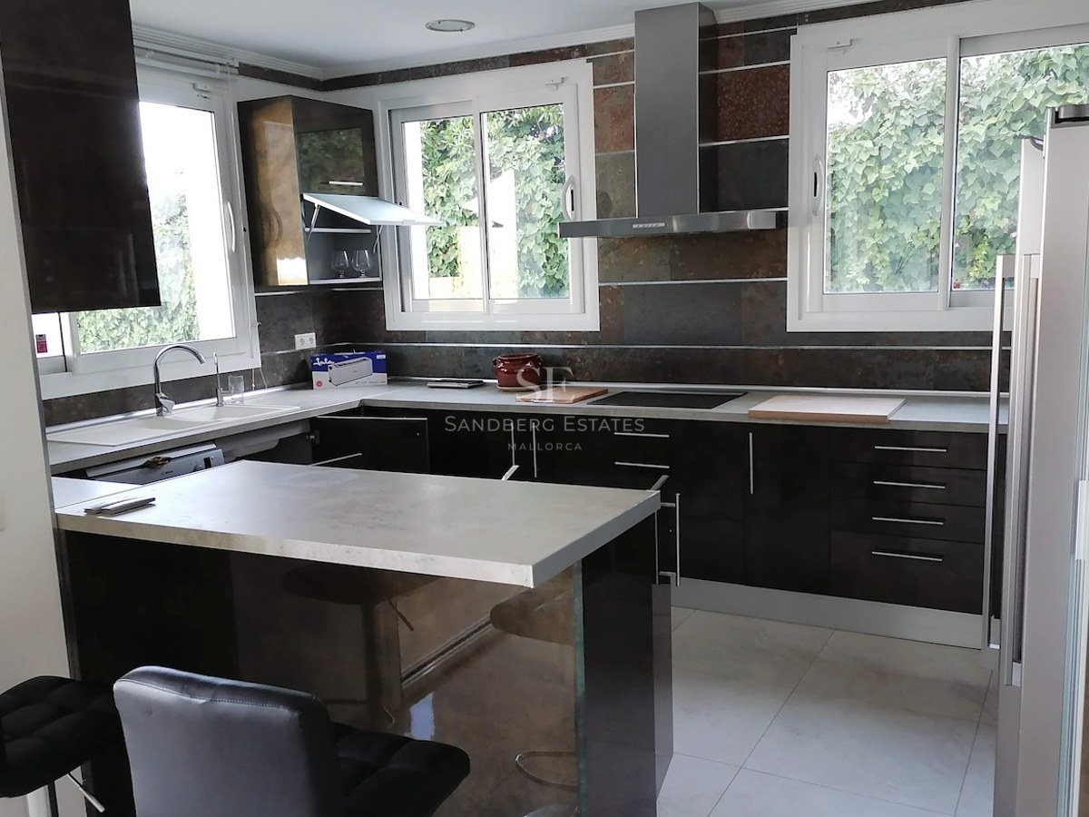 Modern L-shaped kitchen with dark glossy cabinetry, grey countertops, and windows overlooking lush greenery.