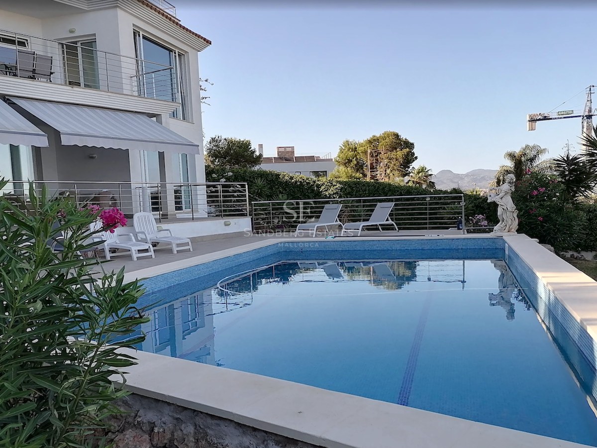 A rectangular blue mosaic-tiled swimming pool in front of a white luxury villa with sun loungers and awnings.