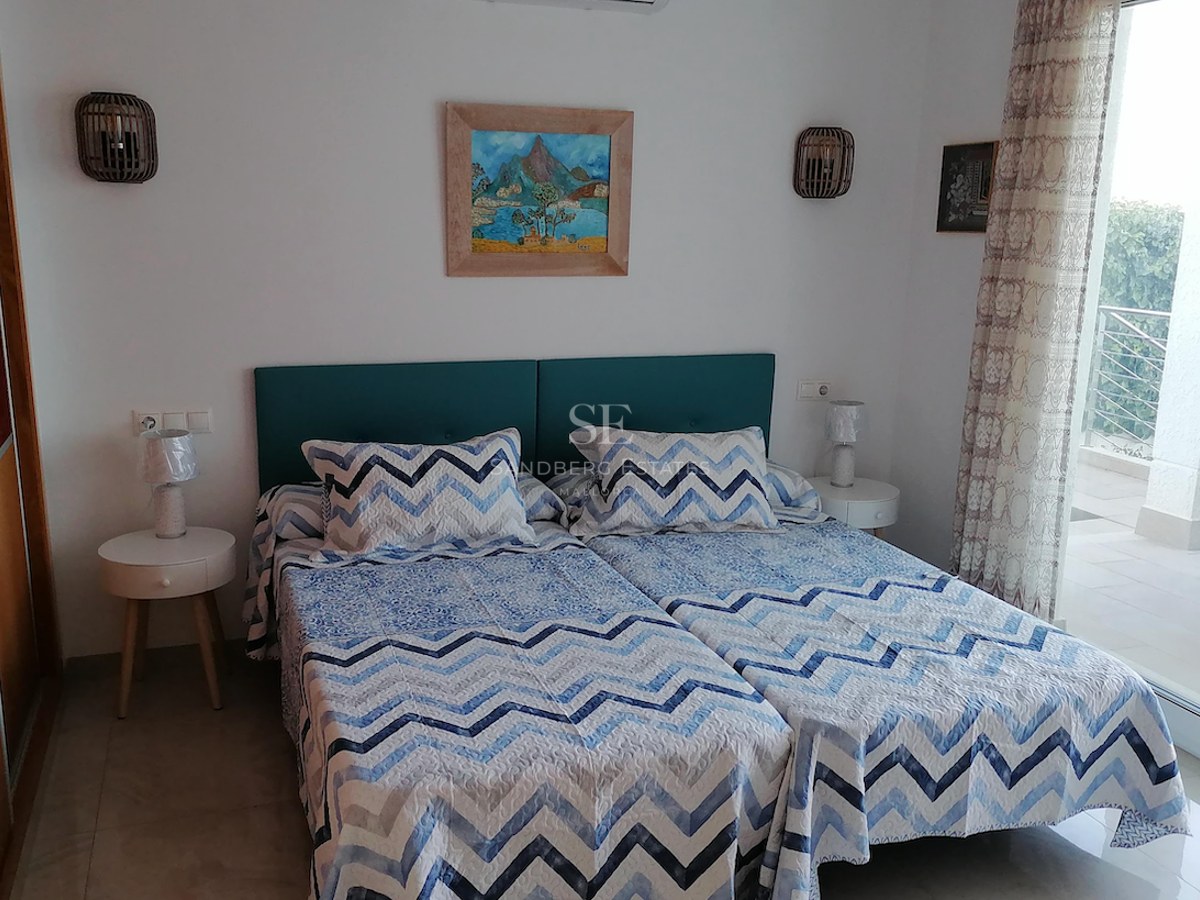Bedroom with twin beds, teal headboard, white walls, and floor-to-ceiling glass door leading to a sunny terrace.