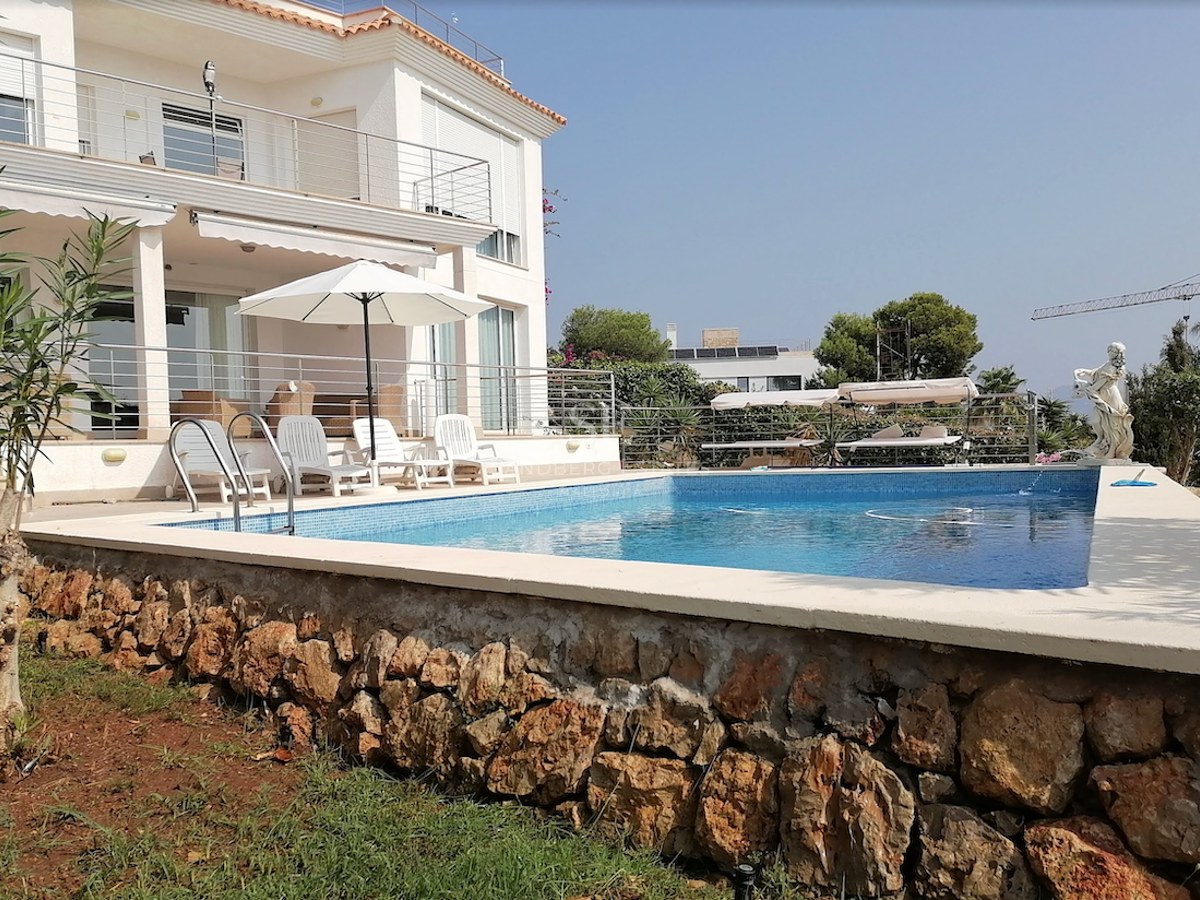 Private swimming pool with lounge chairs and stone retaining wall in front of a white Mediterranean villa.