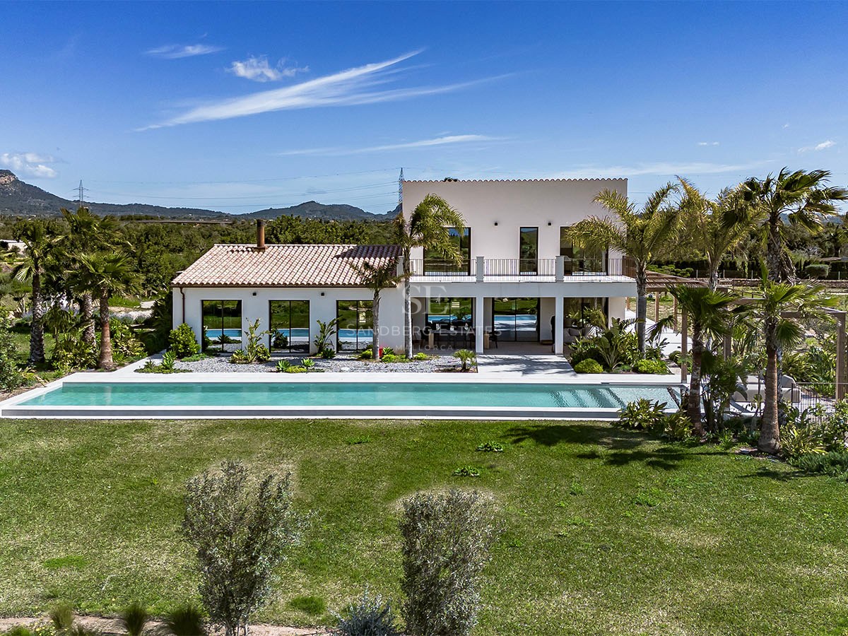 Rear view of a white two-story villa with a long swimming pool, green lawn, and mountain backdrop under a blue sky.