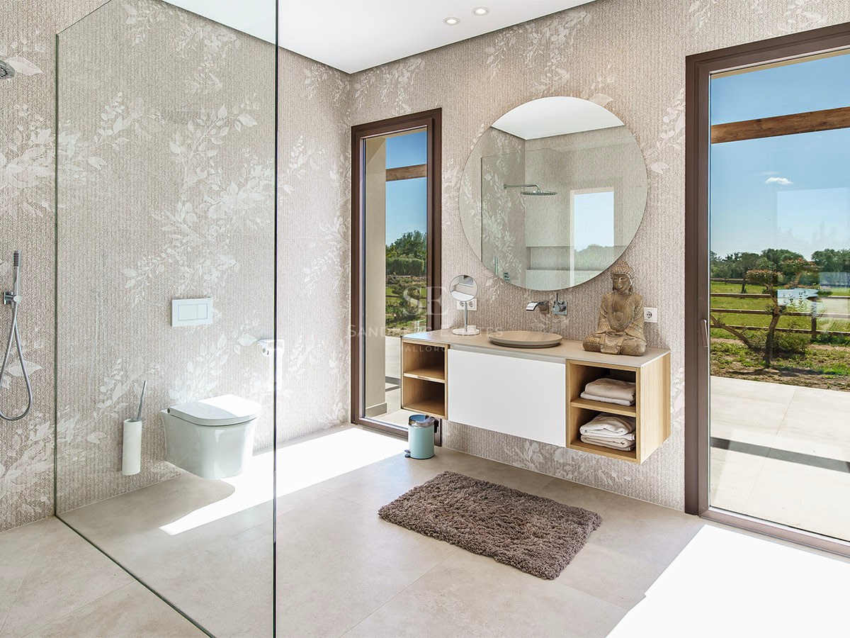Modern bathroom featuring a floating wooden vanity, circular mirror, and floor-to-ceiling windows overlooking a garden.