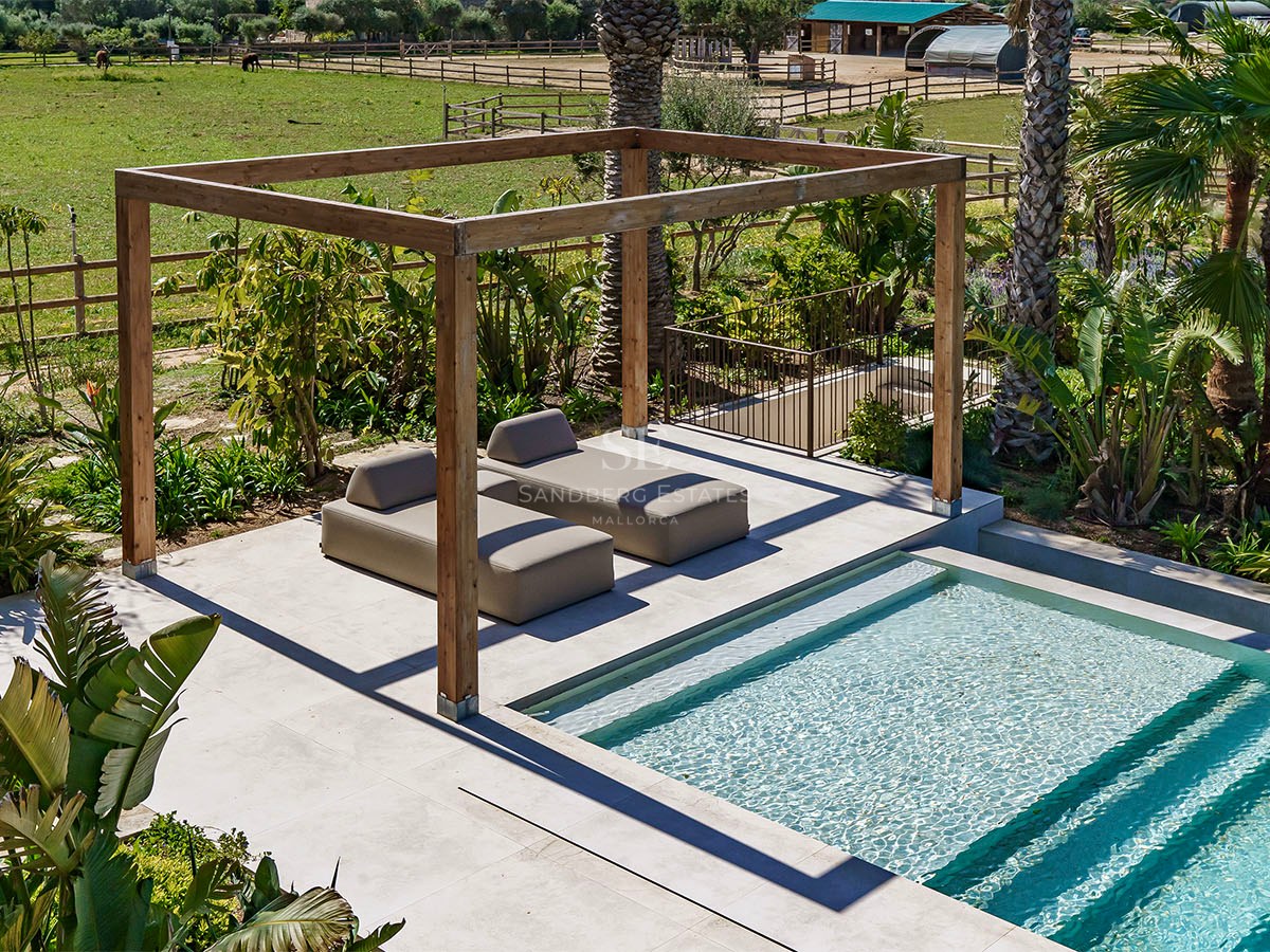 Two modern sunbeds under a wooden pergola beside a turquoise pool overlooking a green field.