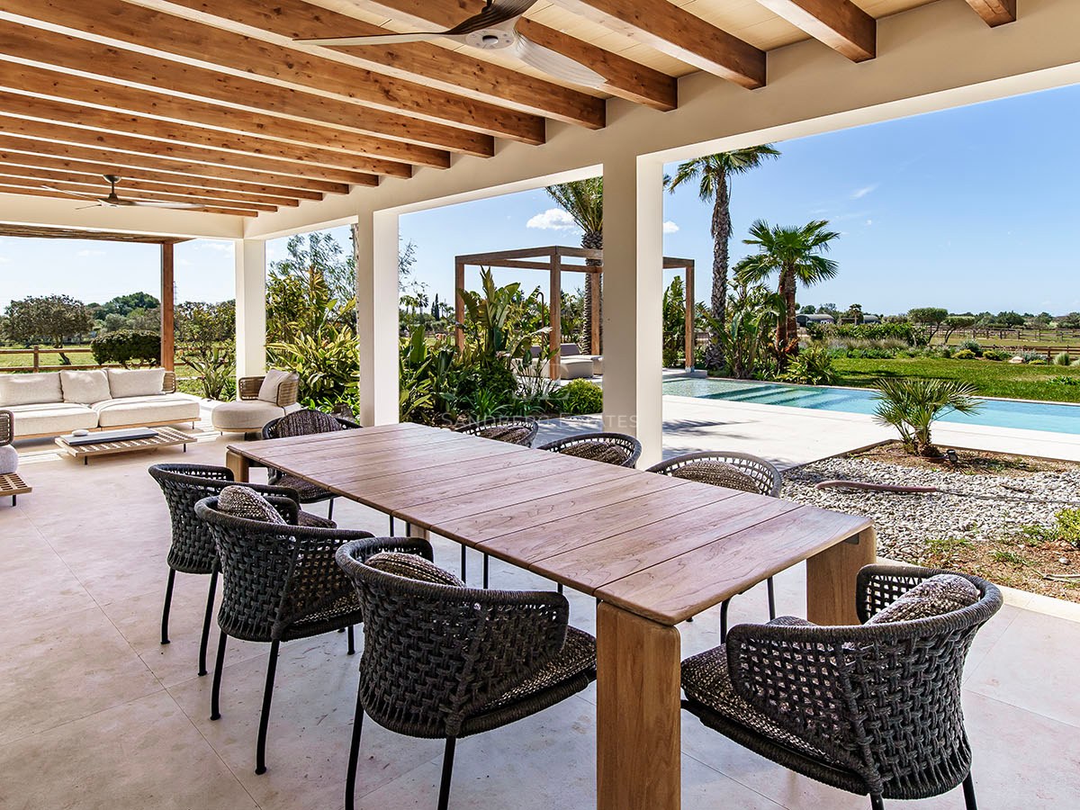 Wooden dining table and woven chairs on a covered terrace with exposed timber beams overlooking a pool and garden.