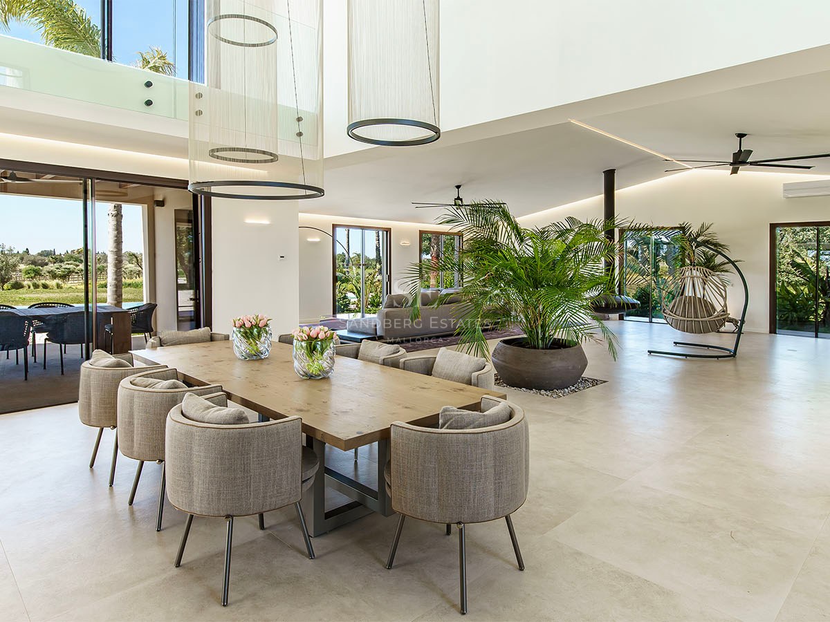 Modern open-plan dining room with a large wooden table, grey chairs, high ceilings, and floor-to-ceiling glass doors.