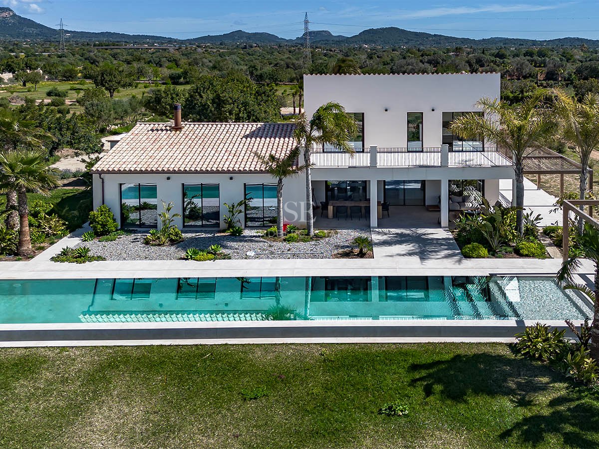 Aerial view of a modern white villa with a turquoise swimming pool and mountains in the background.