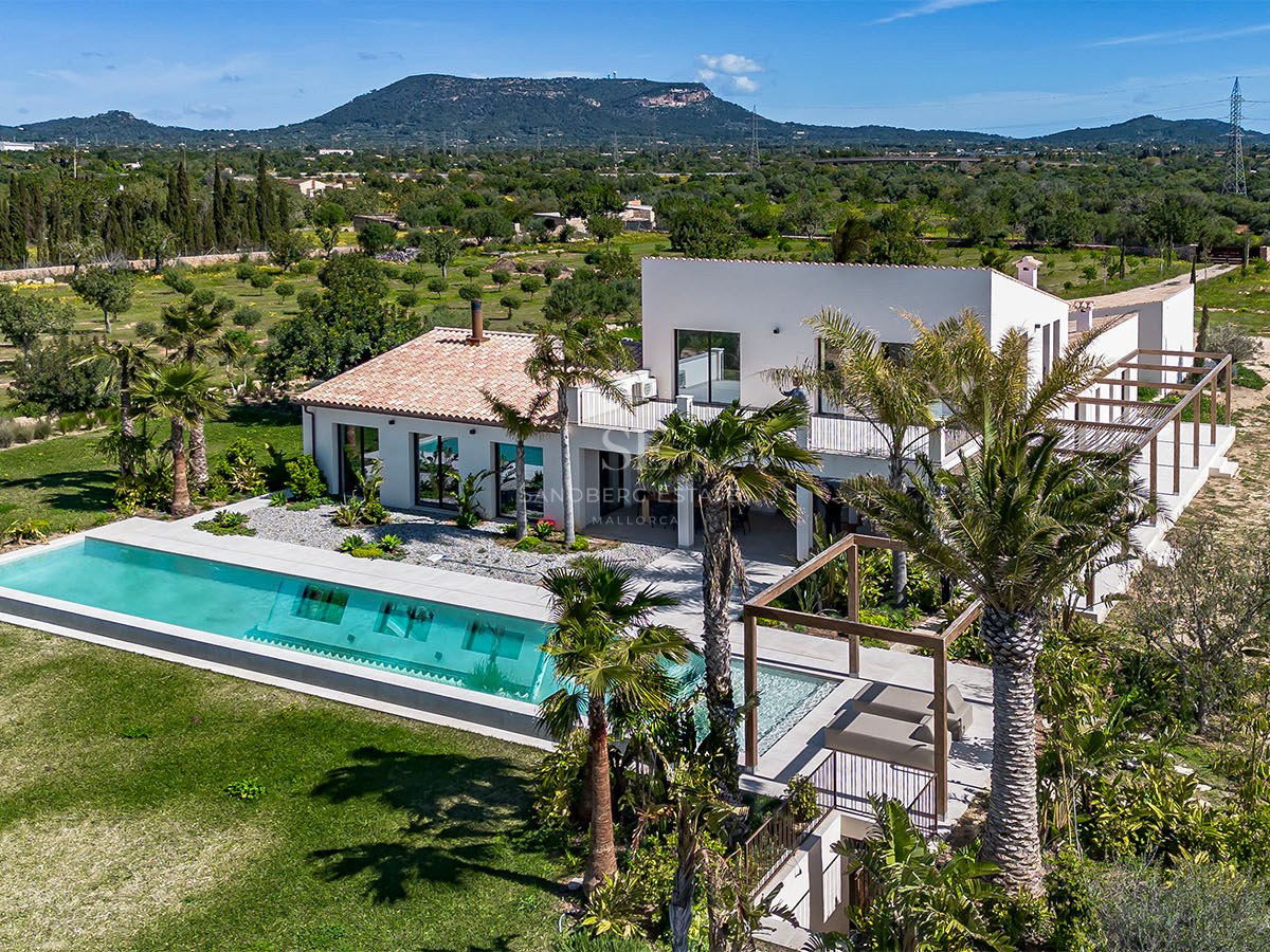 Aerial view of a white luxury villa featuring a long rectangular pool, palm trees, and mountains in the distance.