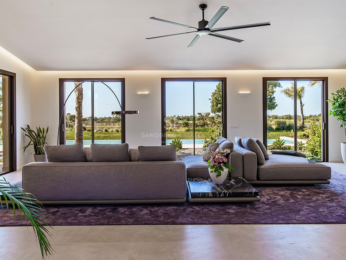 Modern living room with grey sofas, purple rug, and floor-to-ceiling glass doors opening to a lush garden.