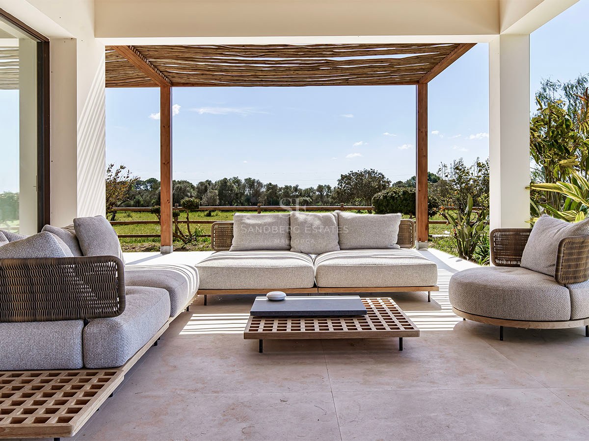 Modern lounge furniture on a shaded stone terrace under a wooden pergola overlooking a Mediterranean landscape.