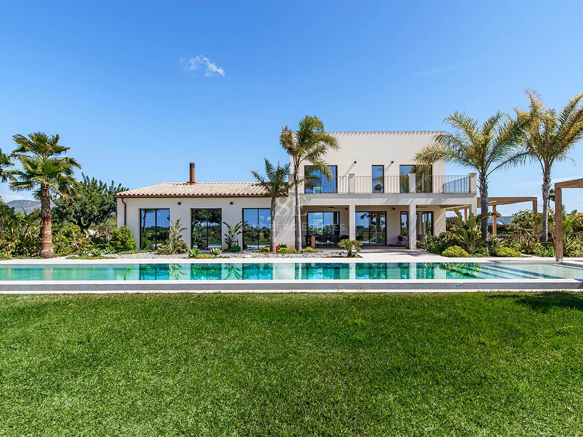 Exterior view of a modern two-story white villa with a large rectangular swimming pool and green lawn under a clear sky.