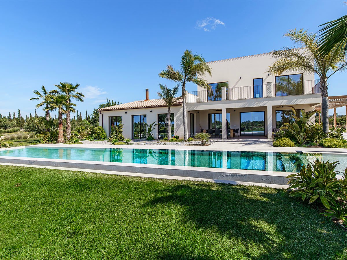 Contemporary white villa featuring a long rectangular swimming pool, manicured green lawn, and palm trees under a clear blue sky.