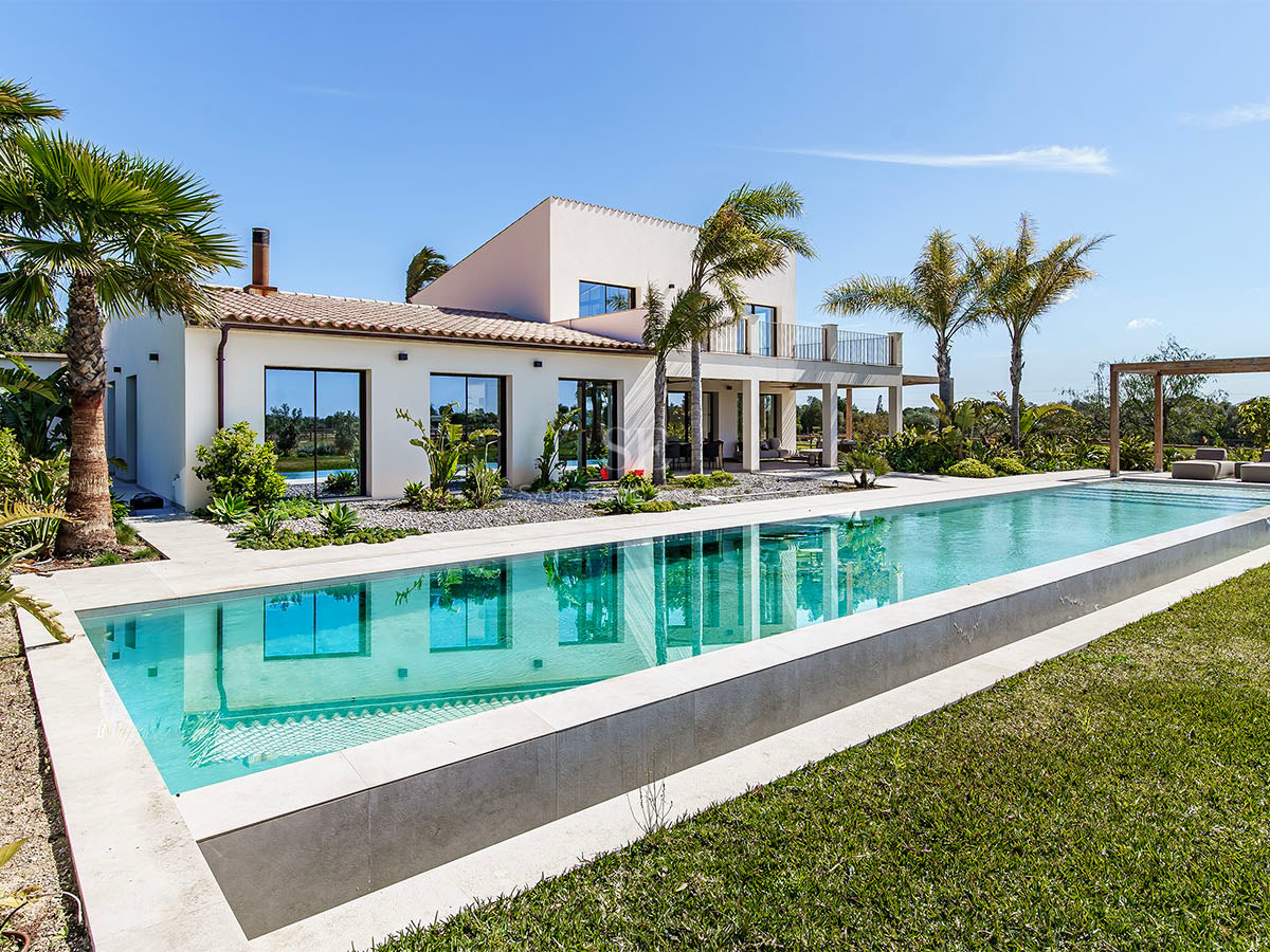 Contemporary white villa with a large rectangular swimming pool, manicured lawn, and palm trees under a clear blue sky.