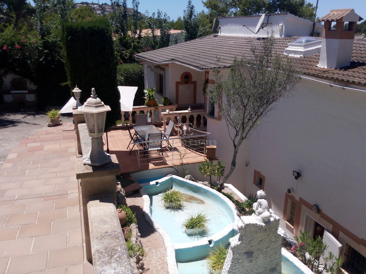 Terrasse en terre cuite avec table à manger en verre, façade blanche et fontaine décorative sous la lumière du jour.