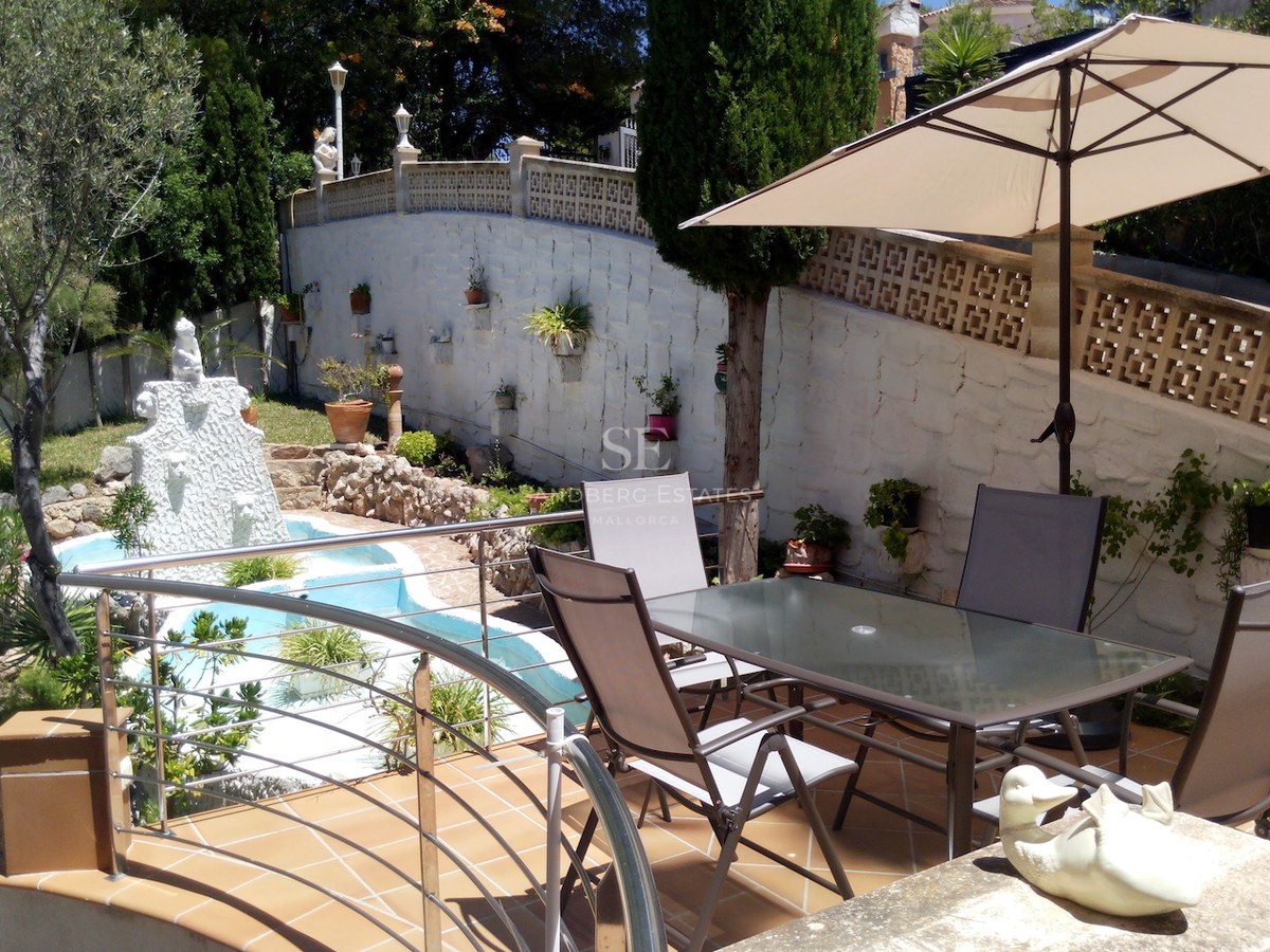 Terrasse avec table en verre et parasol surplombant une fontaine de jardin et des arbres méditerranéens.