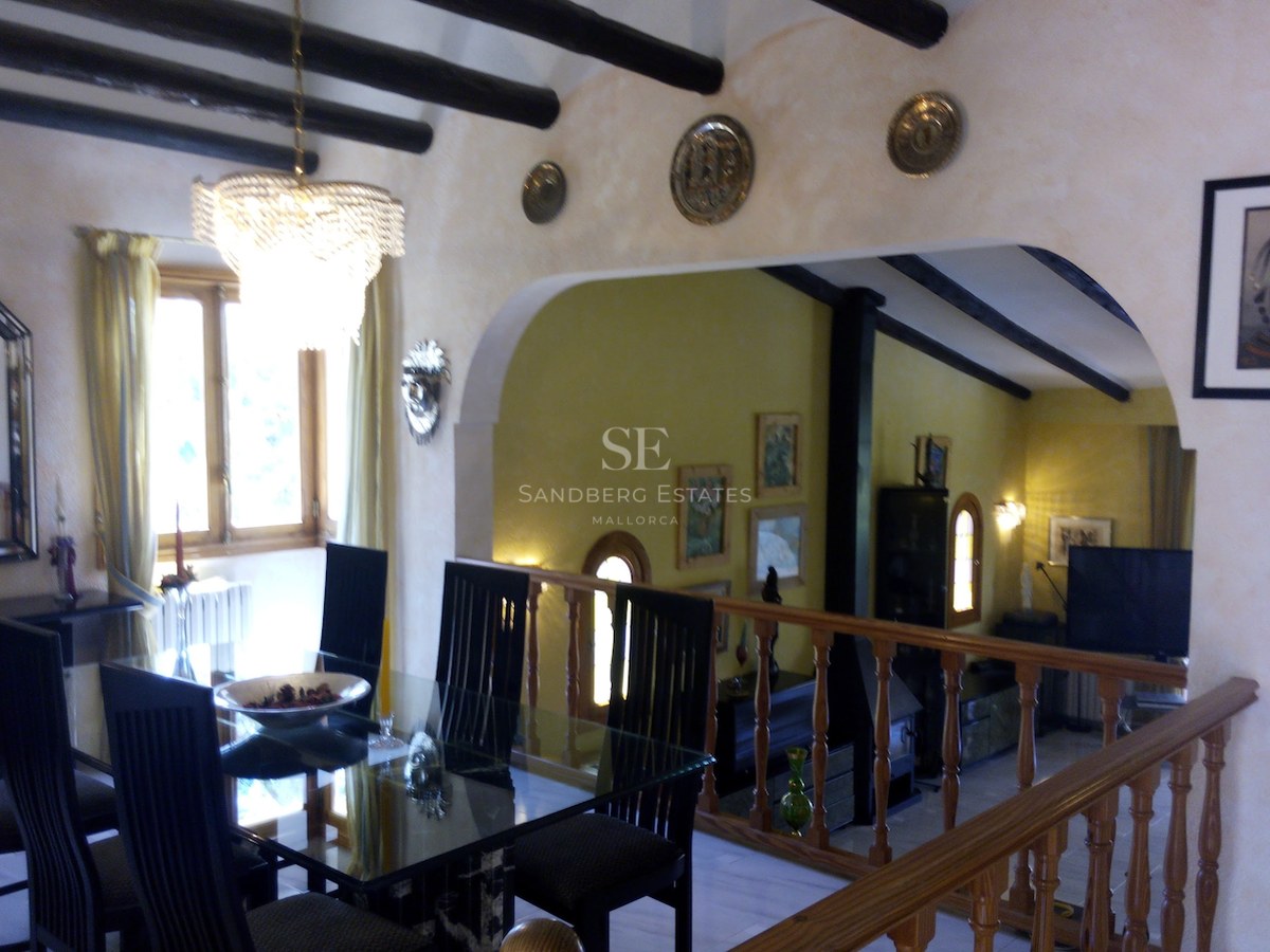 Salle à manger avec table en verre et poutres sombres, avec une balustrade en bois surplombant un salon en contrebas.