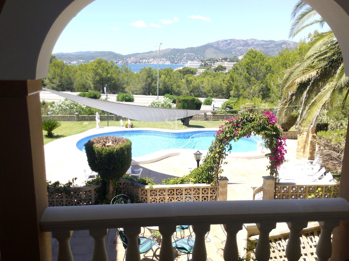 Vue à travers une arche d'une piscine turquoise, d'un jardin luxuriant et de la mer Méditerranée au loin.
