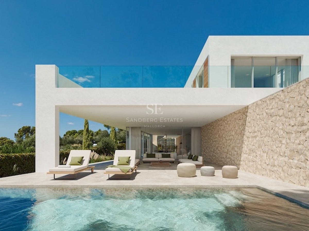 Vy över en pool mot en vit modern villa med skuggad terrass, stenvägg och solstolar under en blå himmel.