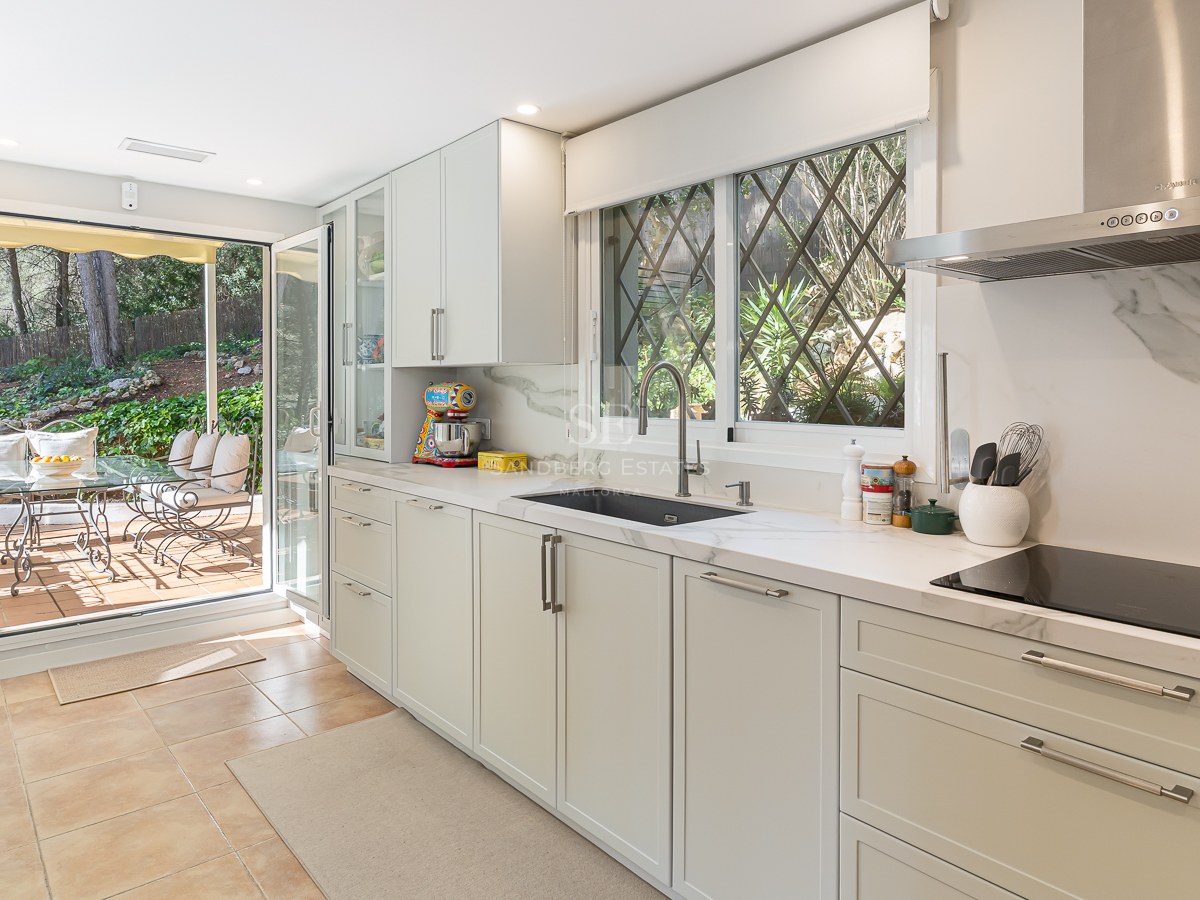 Bright kitchen with white cabinetry, marble countertops, and large glass doors opening to a lush garden terrace.