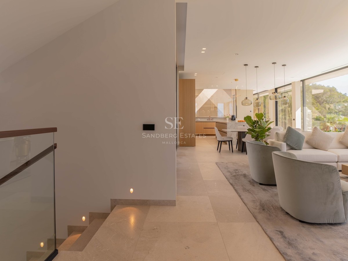 Vue d'un salon moderne à aire ouverte depuis un escalier en pierre avec garde-corps en verre et grandes baies vitrées.