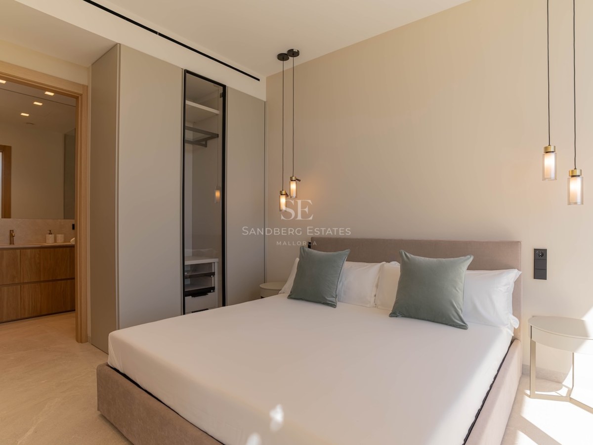 Chambre principale minimaliste avec grand lit, coussins vert sauge, suspensions design et vue sur la salle de bain.