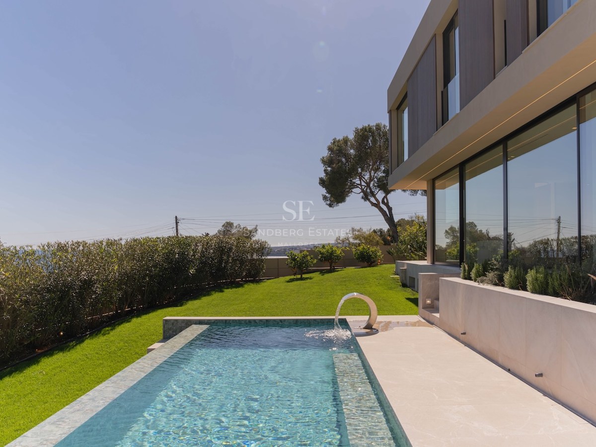 Piscine rectangulaire avec fontaine à côté d'une villa moderne aux grandes baies vitrées et pelouse face à la mer.