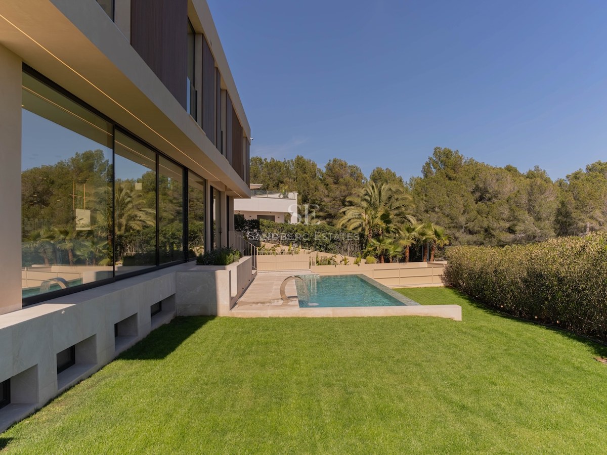 Vue latérale d'une villa moderne avec baies vitrées, piscine rectangulaire et pelouse devant une forêt dense.