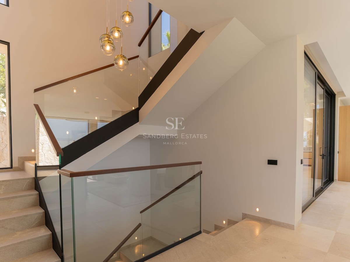Escalier intérieur minimaliste en pierre avec balustrades en verre et suspensions dorées dans une villa lumineuse.