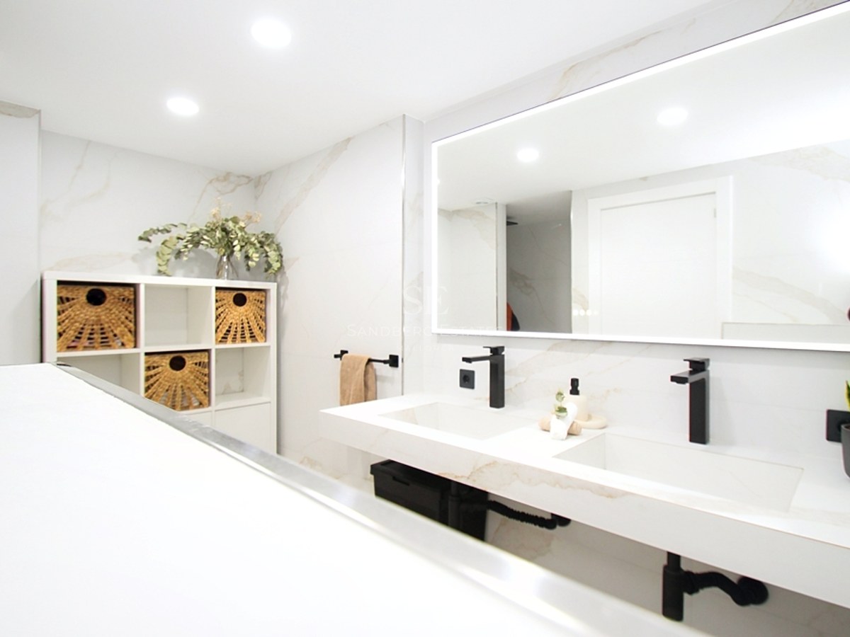 Baño moderno luminoso con lavabo doble de mármol, grifería negra mate y un gran espejo retroiluminado.