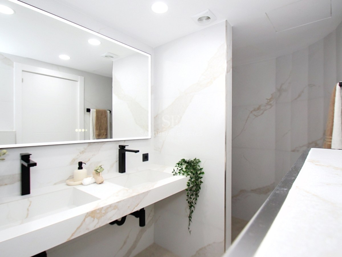 Baño luminoso con lavabo doble de mármol blanco, grifería negra mate y un gran espejo retroiluminado.