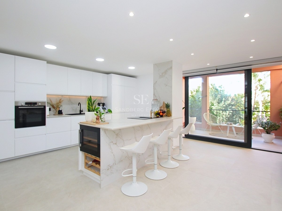 Cocina blanca minimalista con isla de mármol, vinoteca y puertas de cristal que dan a una terraza soleada.