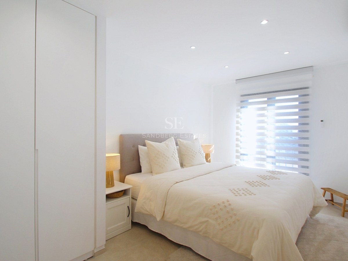 Dormitorio moderno de tonos neutros con cama doble, armarios blancos empotrados y luz natural a través de estores.