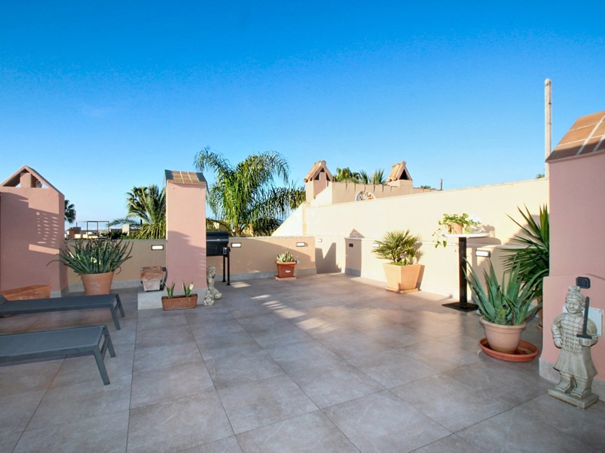 Gran terraza en azotea con azulejos beige, paredes de estuco rosa, tumbonas, plantas y cielo azul despejado.
