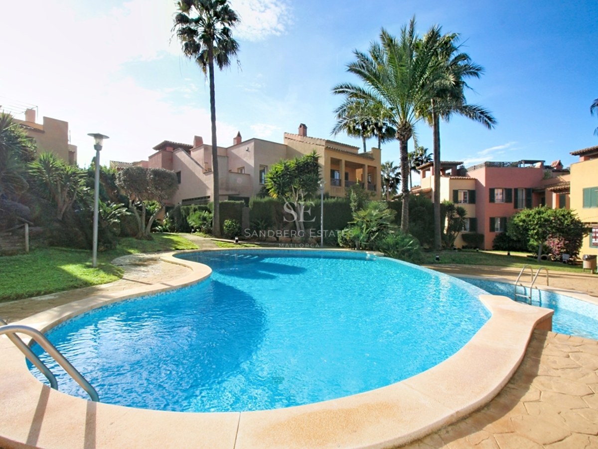 Piscina de forma irregular rodeada de jardines tropicales y edificios mediterráneos bajo un cielo despejado.