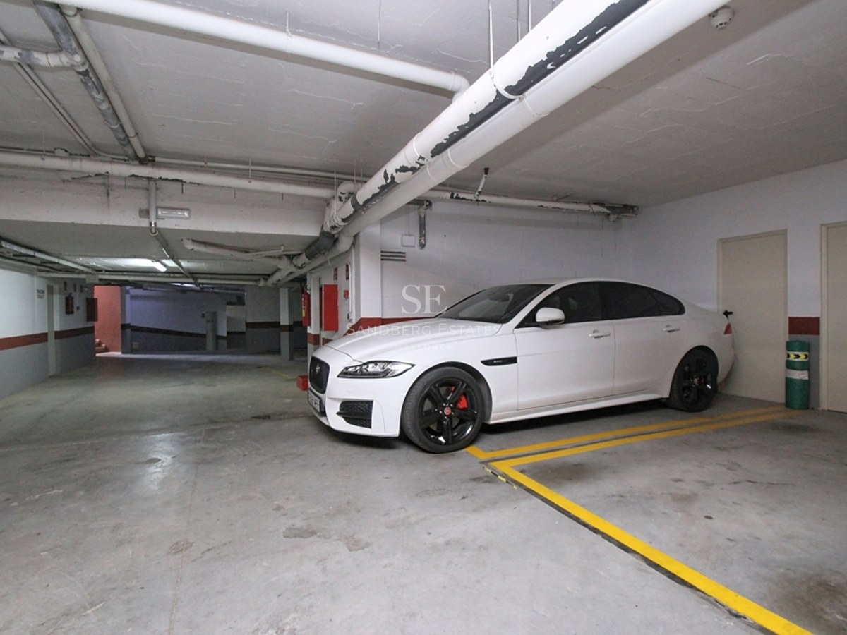 Coche blanco aparcado en un garaje subterráneo iluminado con suelo de hormigón y señalización amarilla.