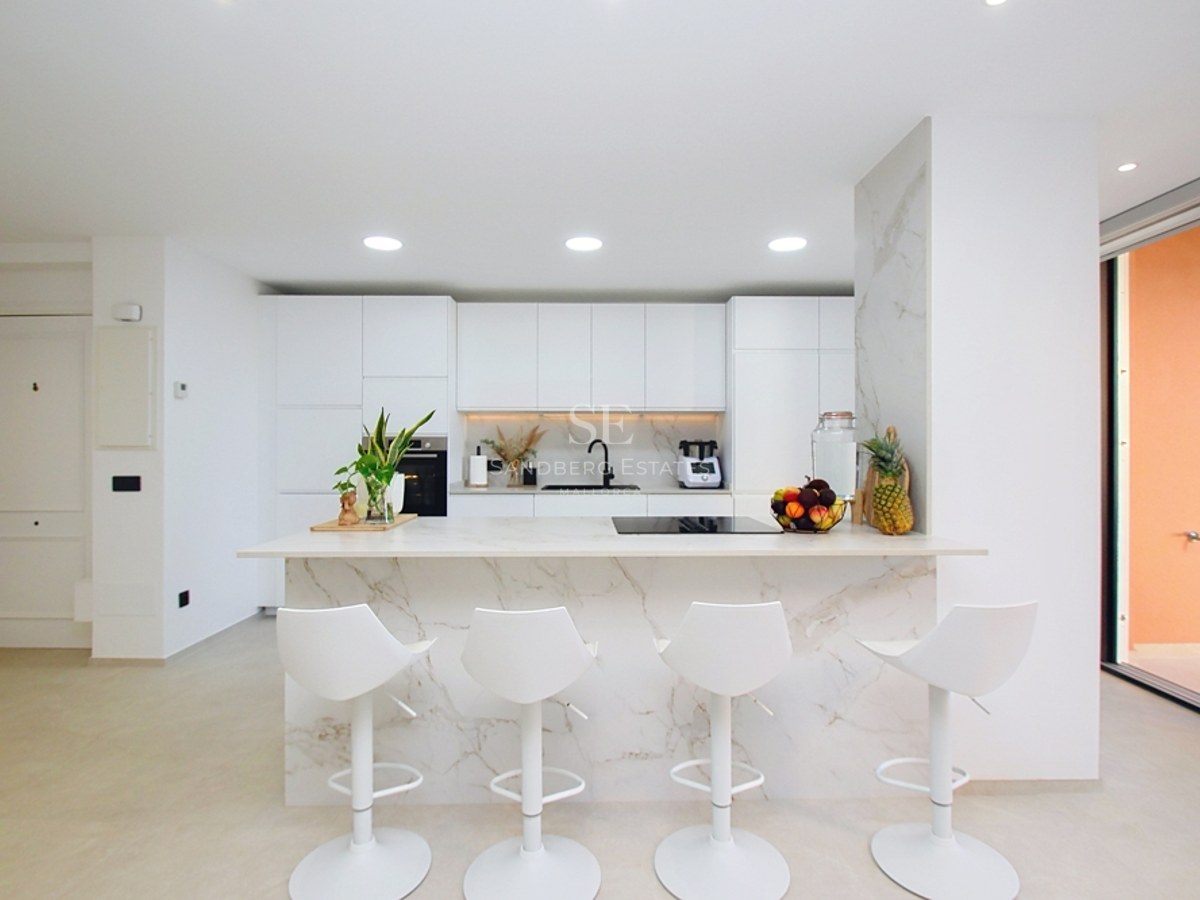 Cocina blanca moderna con una gran isla de mármol, cuatro taburetes blancos y electrodomésticos integrados.
