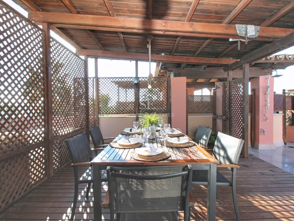 Mesa de comedor bajo pérgola de madera con celosías de privacidad y suelos de tarima en una terraza soleada.