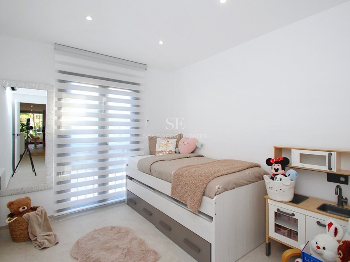 Dormitorio blanco con cama nido, espejo de cuerpo entero, persianas tipo cebra y zona de juegos infantil.