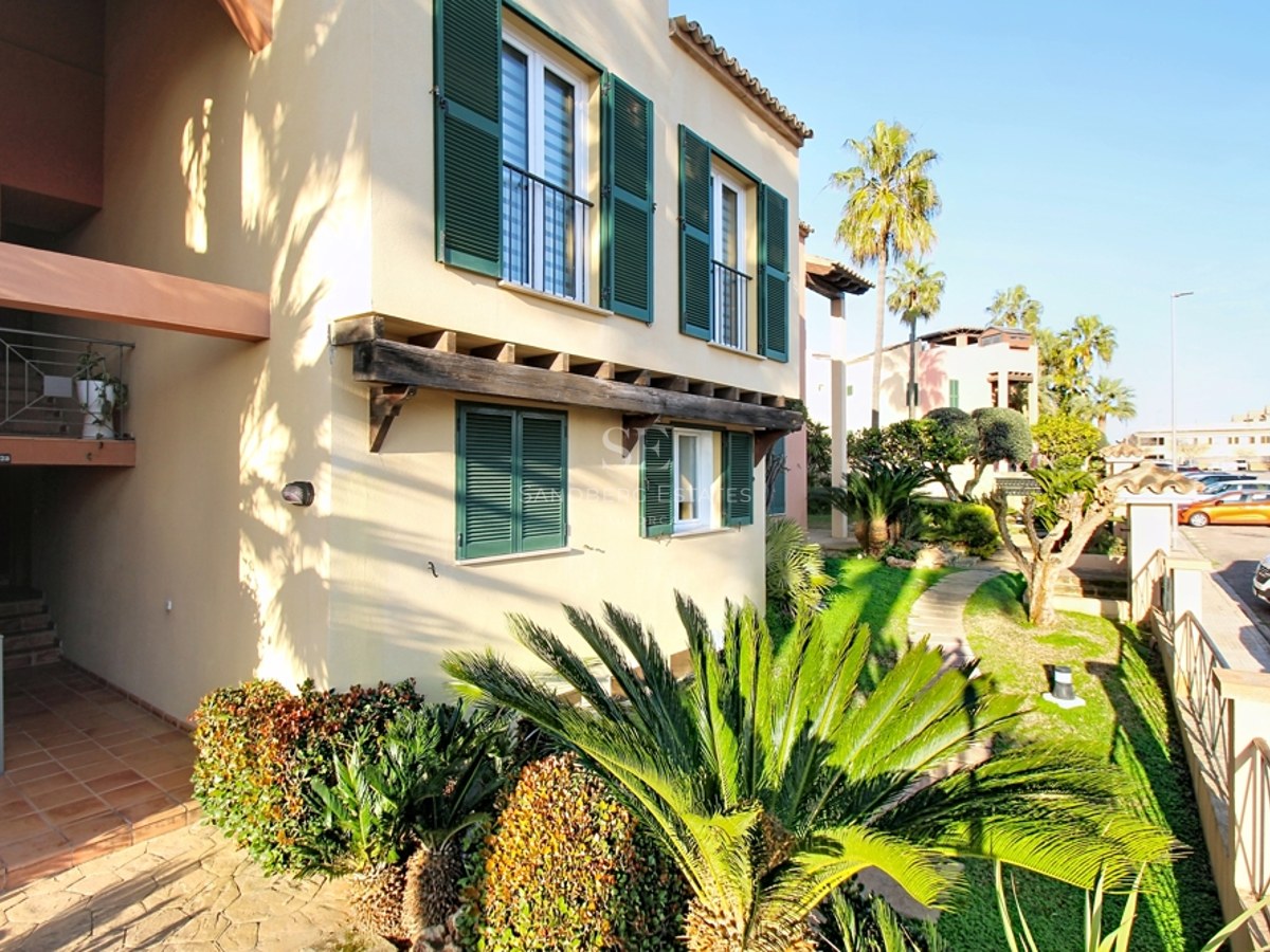 Exterior de edificio beige con persianas verdes, vigas de madera y jardín tropical con palmeras.
