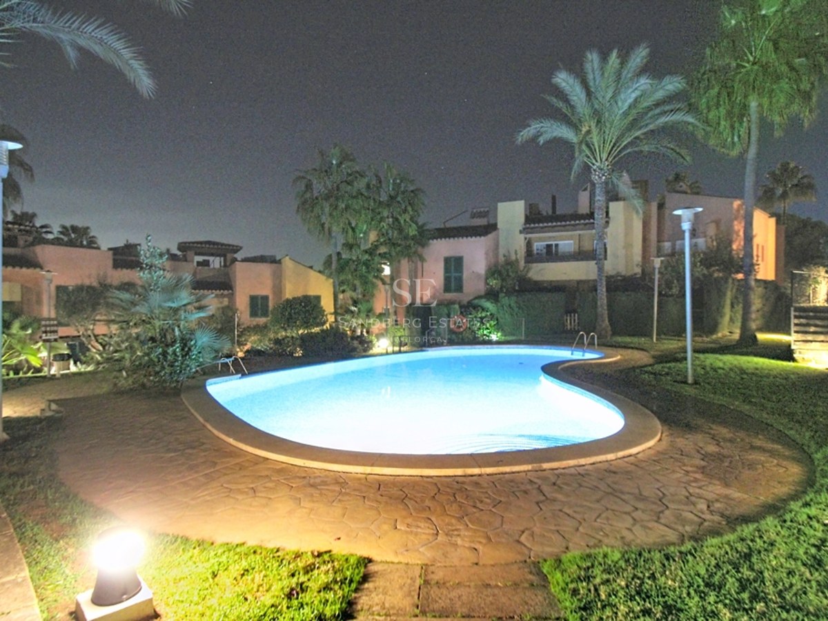 Piscina con forma de riñón iluminada de noche, rodeada de palmeras, césped y villas mediterráneas bajo un cielo oscuro.
